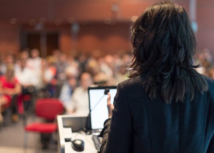 Rear,View,Of,Business,Woman,At,Lectern,Lecturing,At,Conference.